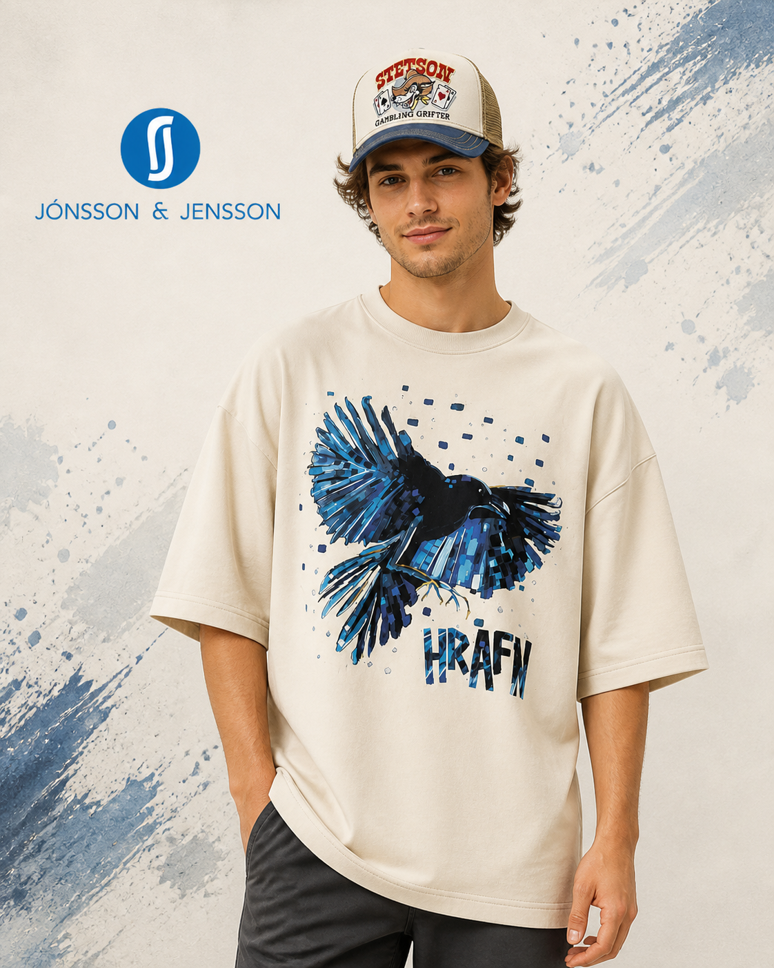 A man wearing a cream-colored oversized t-shirt with a blue raven graphic and the text 'HRAFN'. The background is a textured off-white wall with blue paint splatters.
