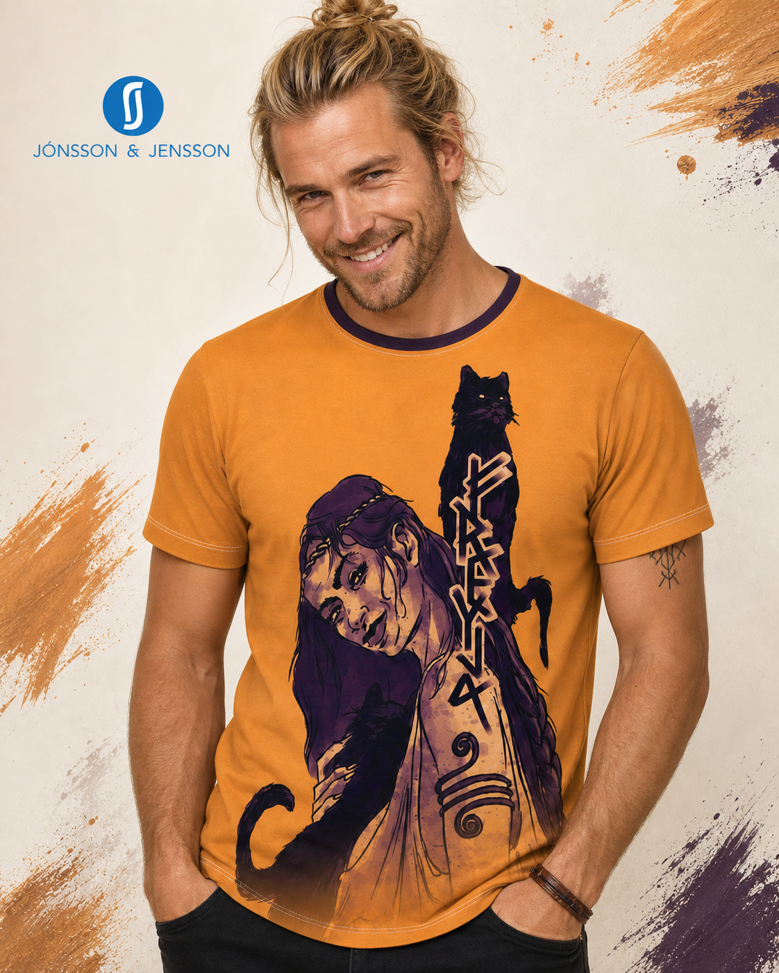 A smiling man with a bun wearing a mustard yellow graphic t-shirt featuring a purple and black illustration of a woman and cat.